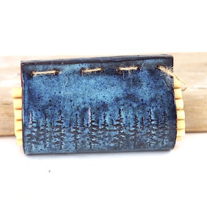 May include: A rectangular, blue ceramic soap dish with a forest silhouette design. The dish has a textured, speckled finish and is accented with natural-coloured wooden slats on the sides. The dish is approximately 10 cm long.