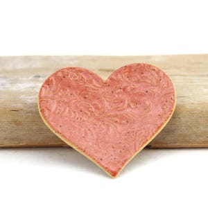May include: A heart-shaped ceramic dish in a dusty rose color featuring an embossed, intricate swirling floral pattern across its surface. The piece has a speckled texture and a smooth, glazed finish with visible light-colored edges.