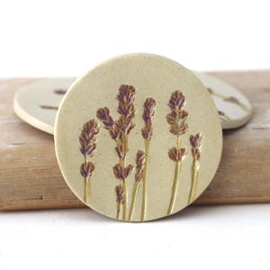 May include: A set of three round ceramic coasters. The coasters are beige with a raised lavender design in shades of brown and gold. The coasters are on a wooden surface.