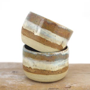May include: Two handmade ceramic bowls stacked. The bowls feature horizontal bands of brown, white, and blue-grey over a beige base. They are displayed on a wooden surface.