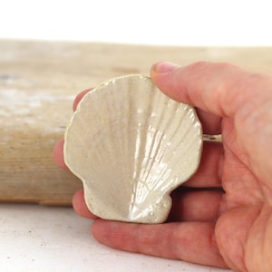 May include: A ceramic seashell-shaped dish in a light beige colour. The dish has a textured surface with radiating lines, and is held in a person's hand. The dish is a decorative item.