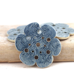 May include: A set of three flower-shaped ceramic buttons in a dusty blue glaze. Each button features a detailed, embossed swirling leaf pattern across the surface and includes multiple circular holes for attachment, with a light-colored base.
