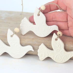 May include: Three white ceramic dove ornaments with a textured floral design, hanging from twine with wooden beads.