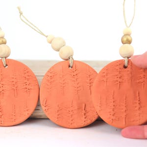 May include: Three terracotta-colored, circular ornaments with a forest design. Each ornament is suspended by a natural cord and adorned with wooden beads. One ornament is being held by a hand.