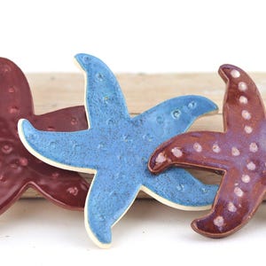 May include: Three ceramic starfish in shades of burgundy and blue. The starfish are arranged on a light-colored wooden plank. The starfish have a glossy finish and are a decorative item.