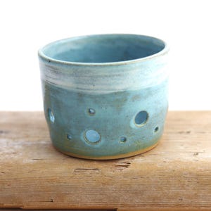 Peut inclure: Un récipient cylindrique en céramique avec un émail bleu clair et une finition lisse. La paroi extérieure présente plusieurs découpes circulaires de tailles variées réparties sur la surface, révélant l'intérieur creux du contenant.