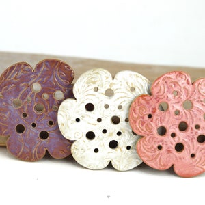 Puede incluir: Tres posavasos de cerámica con forma de flor en morado, blanco y rosa con un diseño floral y agujeros en toda la superficie.