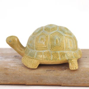 May include: A ceramic turtle figurine featuring a glossy mustard yellow glaze with subtle blue-green accents in the recessed lines of the shell. The turtle has a textured, scaly pattern on its legs and a smooth, rounded head with small dark eyes.
