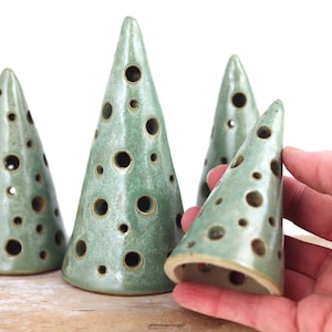 May include: Four ceramic cone-shaped decorations in a seafoam green colour. Each cone has multiple circular openings. One cone is being held by a hand. The cones are displayed on a light brown surface against a white background.