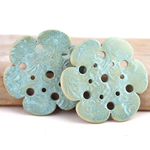 Peut inclure: Deux dessous de verre en céramique en forme de fleur, avec un glaçage marbré bleu clair et turquoise. Chaque dessous de verre comporte une série de trous circulaires et un motif floral détaillé. Les dessous de verre sont présentés sur un morceau de bois flotté.