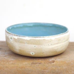 May include: A small, light blue ceramic bowl with a white rim and a speckled beige exterior. The bowl is sitting on a wooden surface.