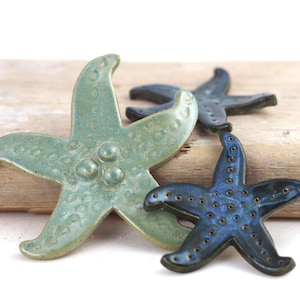 May include: Three ceramic starfish in shades of blue and green rest on a piece of driftwood. The largest starfish is a light teal color with three raised dots. The other two are a darker blue with small dot details.