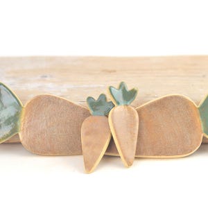 May include: Three ceramic carrot-shaped dishes in different sizes. The dishes are glazed in a light brown colour with green leafy tops.