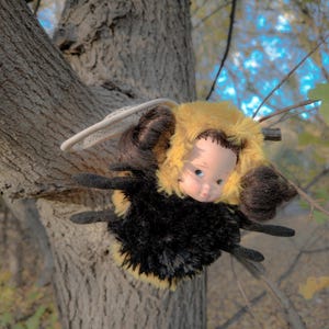 May include: A whimsical handmade doll, resembling a bee, is perched on a tree branch. The doll features a yellow fuzzy head, a doll face with blue eyes, and a black and yellow body. It has dark brown wings and a white halo.