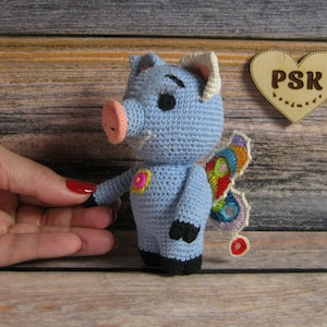 以下が含まれることがあります： カラフルな羽根とピンクの鼻を持つ青い編みぐるみブタ。ブタは「PSK」と書かれた木製のハートのある木製の表面に立っています。