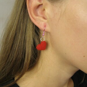 May include: A woman wearing a red heart-shaped dangle earring. The earring is made of crocheted yarn and hangs from a silver hook.
