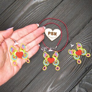 May include: A set of crocheted butterfly jewelry with red hearts. The set includes a necklace and earrings. The necklace is on a red cord and the earrings have silver hooks. The jewelry is on a dark wood surface. The heart on the necklace has the text "PSK" on it.