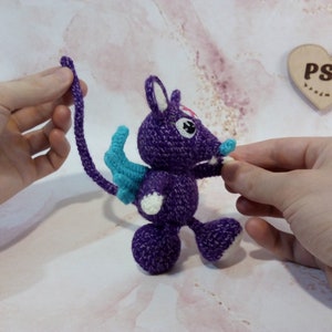 May include: A purple crocheted mouse with blue wings and a long tail. The mouse has white eyes and a pink nose. The mouse is being held by two hands.