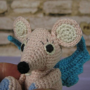 May include: A pink crocheted mouse with blue wings. The mouse has black eyes and a black nose.