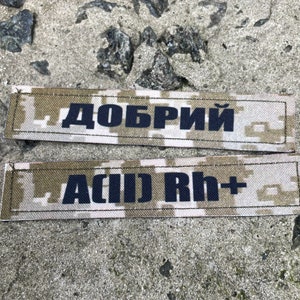 May include: Two rectangular patches with text. The top patch reads "ДОБРИЙ" in dark blue letters. The bottom patch reads "A(II) Rh+" in dark blue letters. Both patches have a camouflage pattern background.