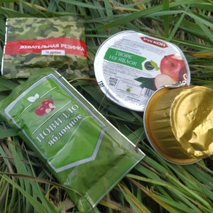 以下が含まれることがあります： 緑と白の袋に「Повидло яблочное」（アップルジャム）の文字と赤いリンゴのイラストが描かれている。緑と迷彩柄の袋に「Жевательная резинка」（ガム）の文字と赤いリンゴのイラストが描かれている。銀色の容器に「Пюре из яблок」（アップルソース）の文字と赤いリンゴのイラストが描かれている。金色のホイル容器。