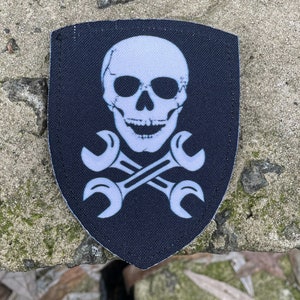 May include: A black patch featuring a white skull and crossed spanners design. The skull has visible teeth and eye sockets. The spanners are positioned beneath the skull, creating a mechanic or industrial theme.