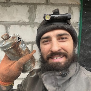 Puede incluir: Un hombre con una boina negra y una linterna frontal sostiene una lata de metal oxidada con un agujero en el lateral. La lata está cubierta de óxido y suciedad.