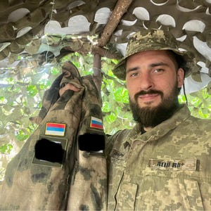 Puede incluir: Una persona con uniforme y sombrero de camuflaje sostiene dos chaquetas de camuflaje. Las chaquetas tienen parches con banderas y rectángulos negros. El fondo incluye redes de camuflaje y follaje.