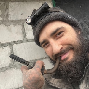 Puede incluir: Un hombre con barba y una linterna en la cabeza sostiene una pipa en la boca. Lleva una chaqueta gris y una boina negra.