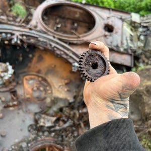 May include: A small, rusty metal gear with many teeth is held up against the backdrop of a destroyed military vehicle. The gear is circular with a central hole, and the vehicle shows signs of fire damage and wear.