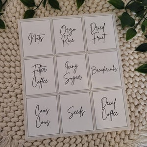 May include: Nine white square labels with black text. The labels are arranged in three rows of three. The text on the labels reads: "Nuts", "Orzo Rice", "Dried Fruit", "Filter Coffee", "Icing Sugar", "Breadcrumbs", "Cous Cous", "Seeds", "Decaf Coffee".