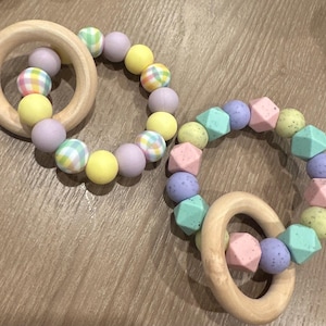 May include: Two wooden teething rings with colorful silicone beads. One ring has yellow, purple, and plaid beads. The other ring has green, purple, and pink beads.
