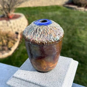 May include: A small, textured ceramic vase with a blue glaze around the opening. The vase has a metallic, iridescent finish.