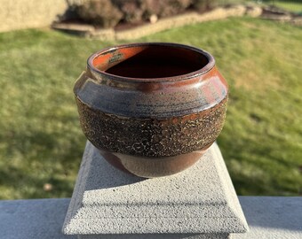 Könnte beinhalten: Ein kleiner, runder, brauner Keramiktopf mit einer strukturierten Oberfläche. Der Topf hat eine dunkelbraune Glasur mit einem Hauch von Rot und Gold.