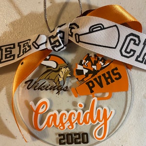May include: A clear round ornament with a white background and orange and black accents. The ornament features a Viking helmet, a megaphone with the text "PVHS" and the text "Cassidy 2020".