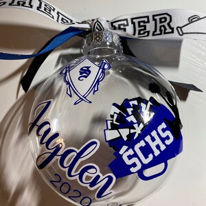 May include: Clear glass ornament with a blue and white cheerleading pom-pom design, the text "SCHS" and the name "Jayden 2020".