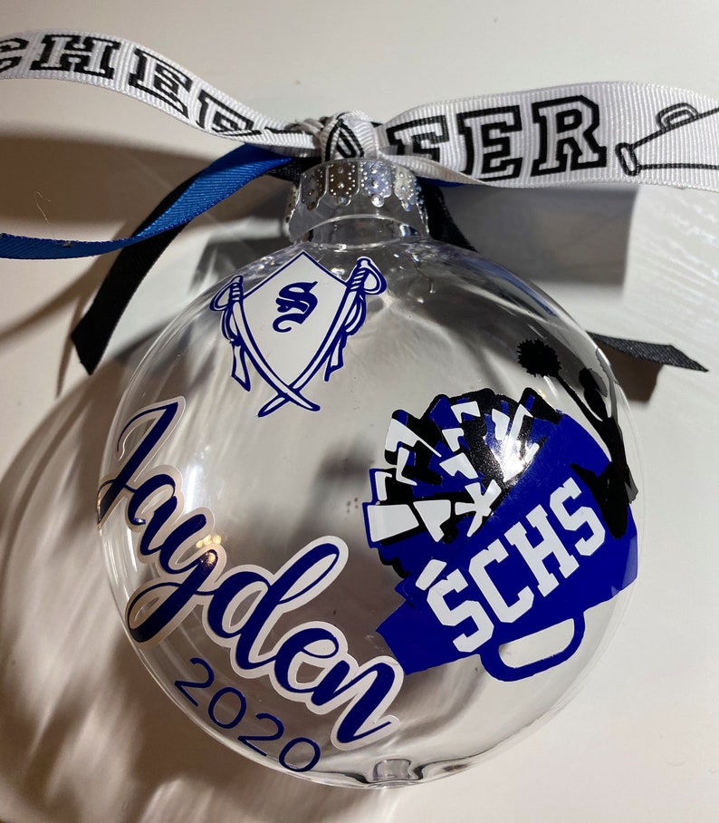 May include: Clear glass ornament with a blue and white cheerleading pom-pom design, the text "SCHS" and the name "Jayden 2020".