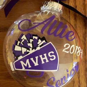 May include: A clear glass ornament with a purple and white megaphone and pom pom design. The ornament also has the text "Allie", "MVHS", "2018-19", and "Senior" written on it.
