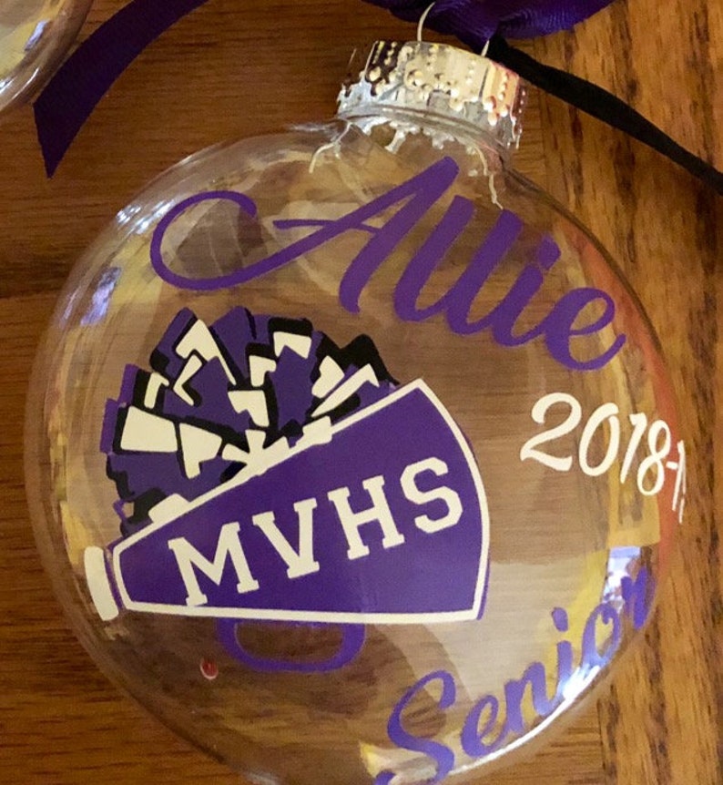 May include: A clear glass ornament with a purple and white megaphone and pom pom design. The ornament also has the text "Allie", "MVHS", "2018-19", and "Senior" written on it.