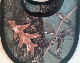 May include: Camouflage fabric baby bib with a black trim and the words "Burly Camo" printed on the bottom right corner.