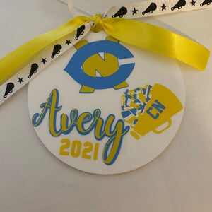May include: A white round ornament with a blue and yellow design. The ornament has the name Avery and the year 2021 written on it. The ornament is decorated with a blue and yellow letter C and a yellow and blue cheerleader pom-pom.
