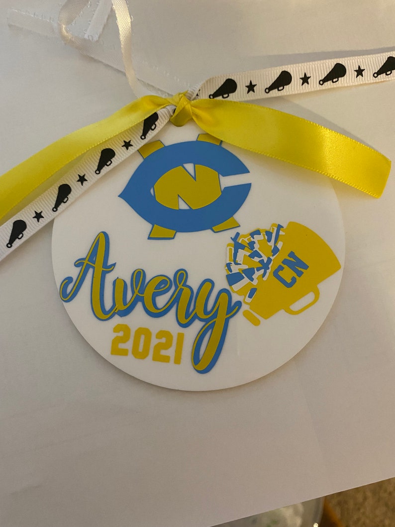 May include: A white round ornament with a blue and yellow design. The ornament has the name Avery and the year 2021 written on it. The ornament is decorated with a blue and yellow letter C and a yellow and blue cheerleader pom-pom.