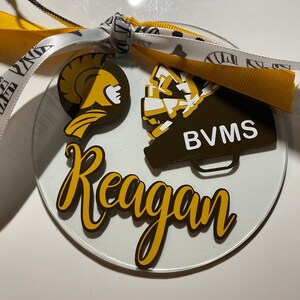 May include: A clear glass ornament with a gold and white design. The ornament features a Spartan helmet, a megaphone with the text "BVMS", and the name "Reagan".