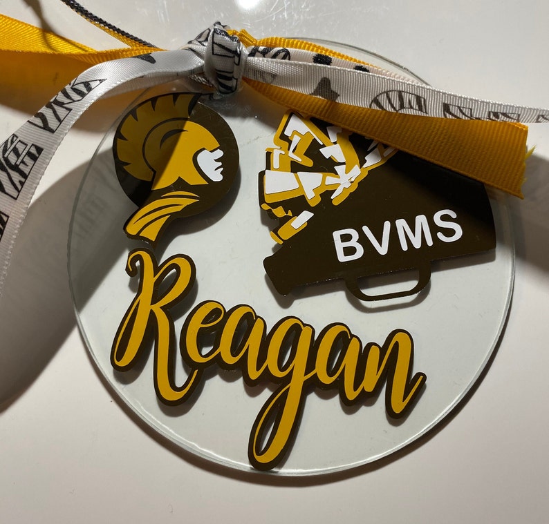 May include: A clear glass ornament with a gold and white design. The ornament features a Spartan helmet, a megaphone with the text "BVMS", and the name "Reagan".
