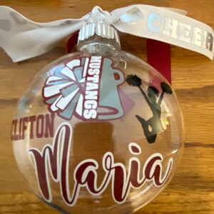 May include: Clear glass Christmas ornament with a maroon and white cheerleading pom pom, a maroon and white cheerleader silhouette, a maroon and white "Mustangs" logo, and the name "Maria" in maroon cursive writing.