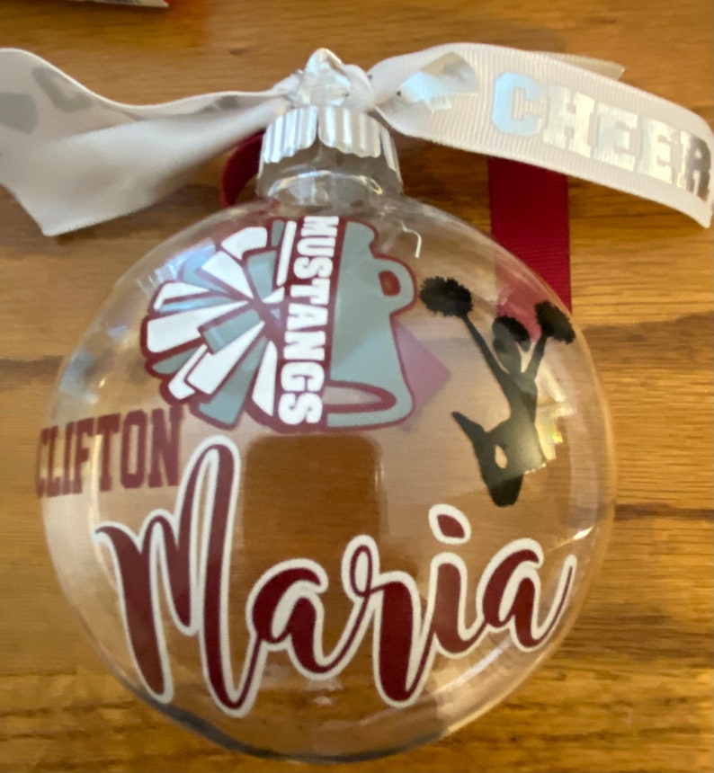 May include: Clear glass Christmas ornament with a maroon and white cheerleading pom pom, a maroon and white cheerleader silhouette, a maroon and white "Mustangs" logo, and the name "Maria" in maroon cursive writing.