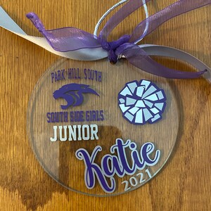 May include: A clear round ornament with purple text and a white and silver cheerleading pom-pom. The text reads "Park Hill South South Side Girls Junior Katie 2021".