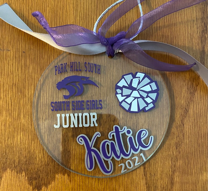 May include: A clear round ornament with purple text and a white and silver cheerleading pom-pom. The text reads "Park Hill South South Side Girls Junior Katie 2021".