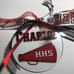 May include: A white round glass ornament with a red and black megaphone, the text "HHS" and the name "CHARLIE" in black and red lettering. The ornament is decorated with red and black ribbons.