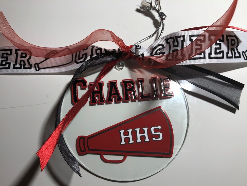 May include: A white round glass ornament with a red and black megaphone, the text "HHS" and the name "CHARLIE" in black and red lettering. The ornament is decorated with red and black ribbons.